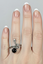Load image into Gallery viewer, A close-up of a person's hand wearing a sterling silver ring with a black onyx gemstone oval stone, with a display tray of rings in the background on a wooden table.
