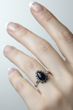 Load image into Gallery viewer, wearing a sterling silver ring with a black onyx teardrop-shaped stone. In the background, a display tray of rings sits on a wooden table next to a laptop.
