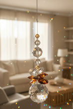 Load image into Gallery viewer, A hanging crystal suncatcher with faceted clear glass beads and a large clear round crystal ball at the bottom, accented by amber-brown.  shown against a plain indoor wall.
