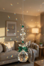 Load image into Gallery viewer, A hanging crystal suncatcher ornament with a faceted crystal ball, green crystal  and clear crystal beads on a thin metal string, shown against an indoor wall.
